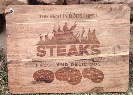 Holztafel mit der Aufschrift "Steaks - frisch und lecker" und dekorativen Fleischmotiven.