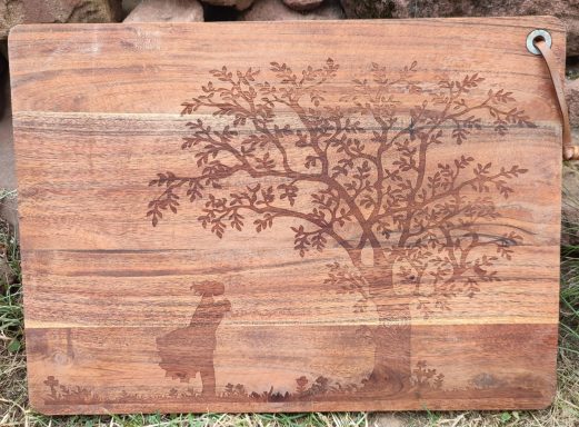 Holzbrett mit eingraviertem Baum und Silhouette eines Hundes im Vordergrund.